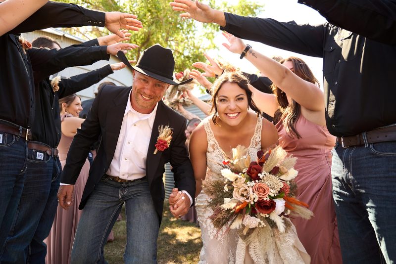 Bride and groom laugh as they run through a tunnel of cheering guests, groom in a black cowboy hat, bride holding a fall bouquet