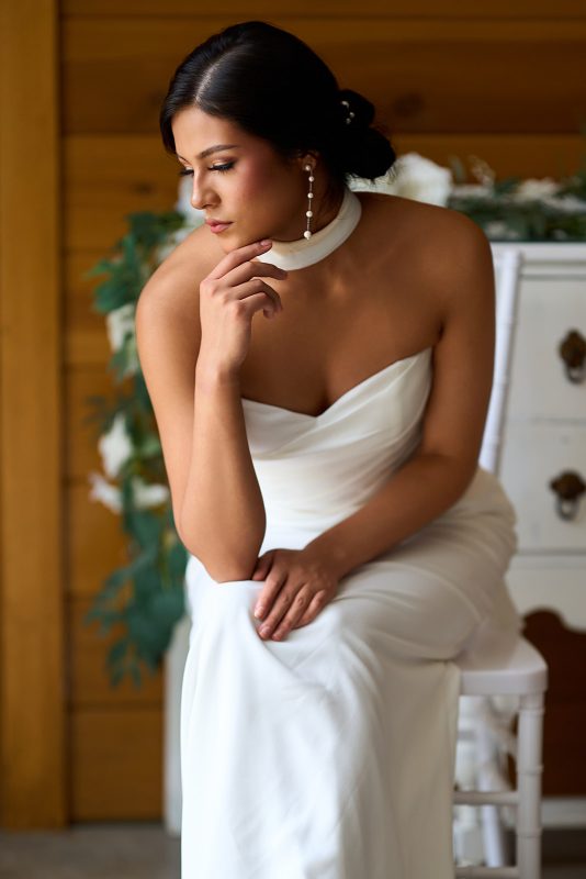 Seated bridal portrait — bride in a strapless white gown and pearl drop earrings, chin resting on her hand