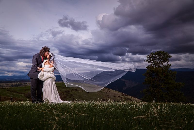 A couple kissing under storm clouds, her veil lifting in the wind