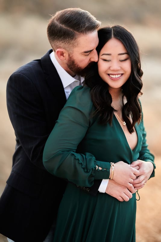 A man kissing his fiancée on the cheek as she smiles, in a green dress in a golden field
