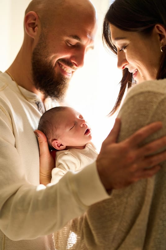 A mother and father smiling down at their sleeping newborn, held against his chest in soft window light