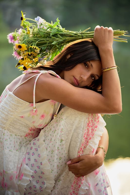 A senior in a floral dress resting her head on her arm, an armful of wildflowers over her shoulder